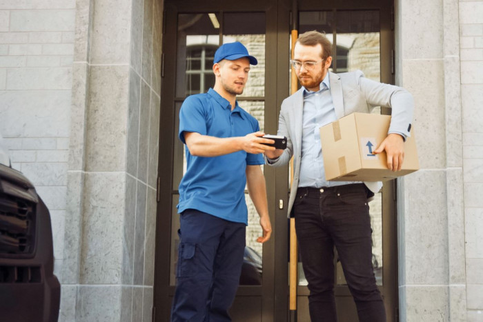 A man holds a large parcel in one hand and makes an electronic signature with the other hand on a device held by a delivery driver 