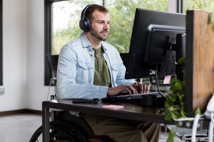A man in casual clothes and headphones uses a computer at a desk in a home office. He  sits in a wheelchair 