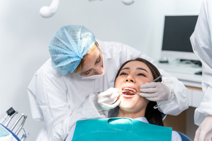 A female dentist wearing hair covering, gloves and a white gown bends over a patient and inserts dental tools into her mouth