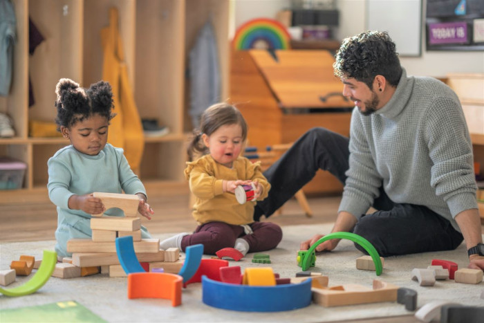 Male Early Childhood Education teacher and two toddlers sit on the floor in an early childhood centre playing with blocks