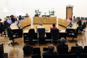A group of people sit in a round circle of desks in Parliament at a select committee hearing