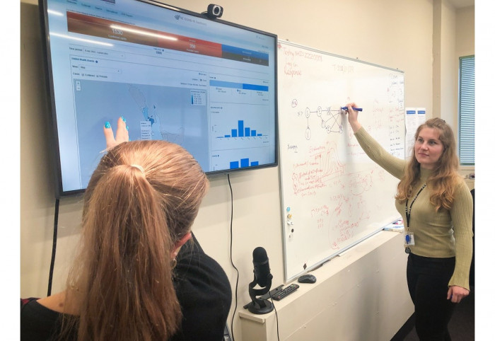 A woman looking at a NZ map on a screen while another woman writes on a whiteboard 