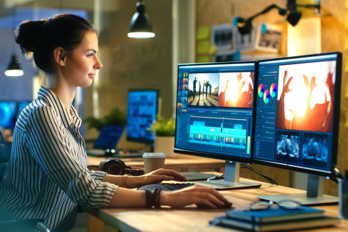 A woman sits at a computer with two screens which show video editing software