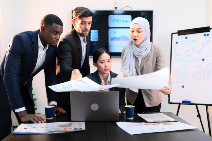 A mixed group of people in business clothes looking at a laptop and some paper reports