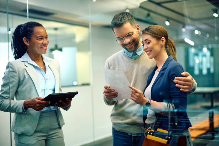 A woman in a business suit holds a folder looking at a couple who are smiling and looking at a piece of paper in an office 