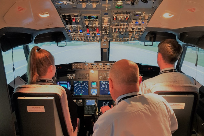 Two students seated in the flight deck of a flight simulator, with an instructor behind them