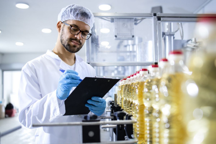 A man in a hair net and a lab coat and gloves writes on a clipboard as bottles of oil run past him on a  conveyor belt