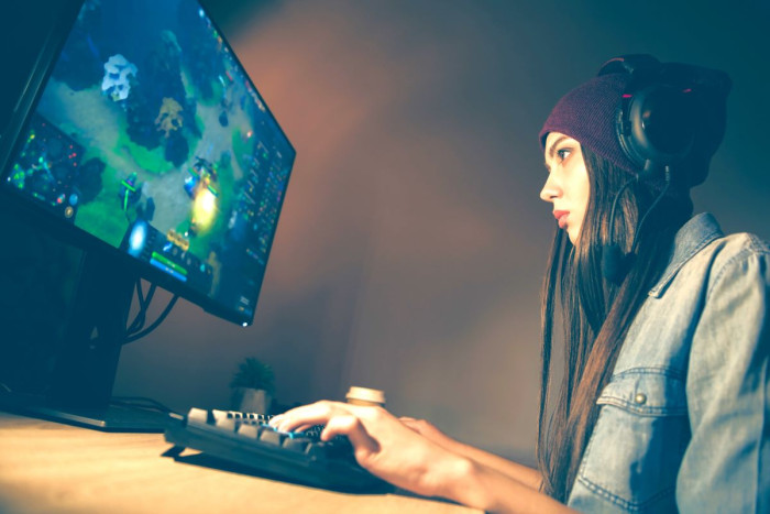 A woman wearing headphones sits at a table playing a video game on a large computer screen 
