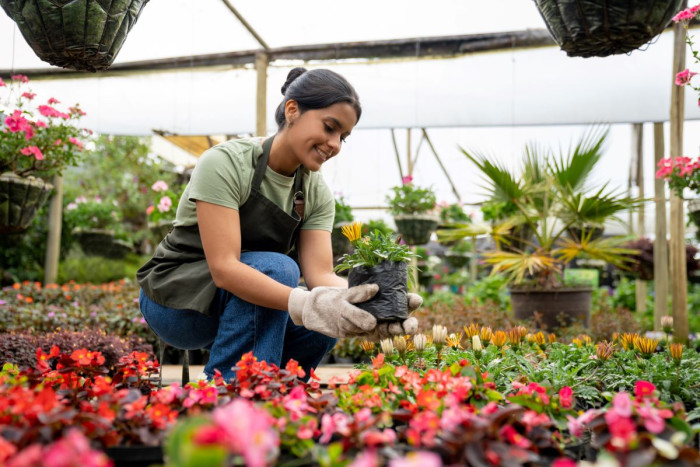 A smiling woman wearing gardening gloves is placing a pot in a display of flowers 