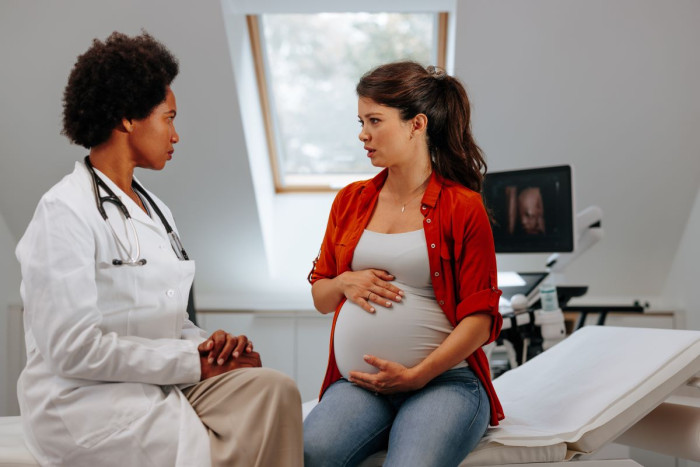 A pregnant person sits in a doctor's rooms talking to a doctor