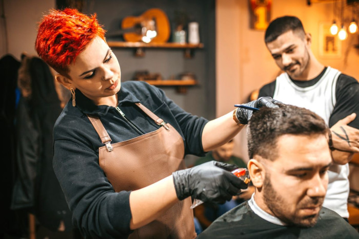 A young woman with bright red hair wearing an apron clips the hair on the back of a man's head while another man looks on 