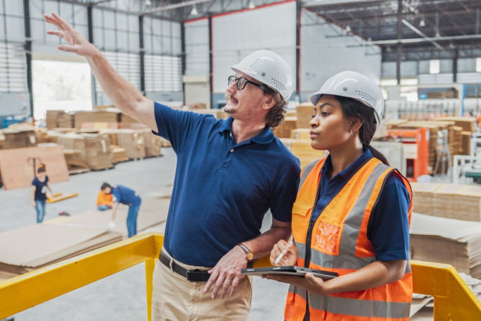 An older man and a younger woman in a high vis vest stand above a factory. She has a clipboard 