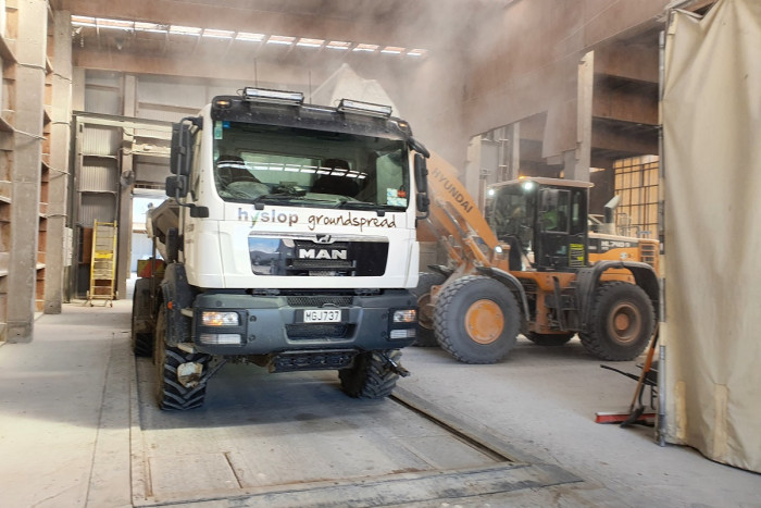 A truck is parked inside a dusty warehouse. Another machine is loading it with fertiliser 