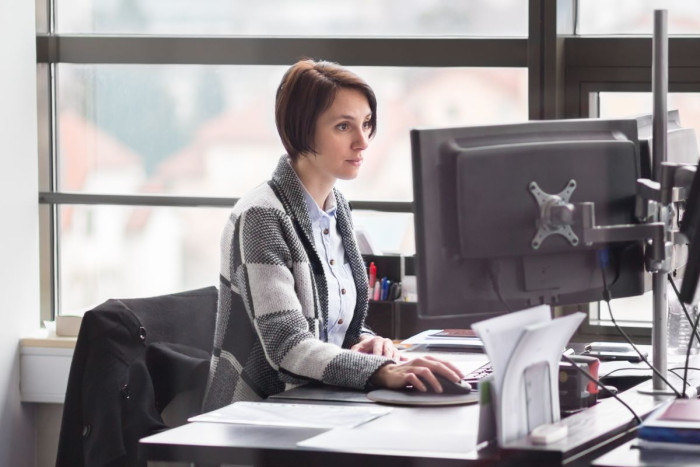 A woman in business clothes sits at an office desk using a desktop computer 