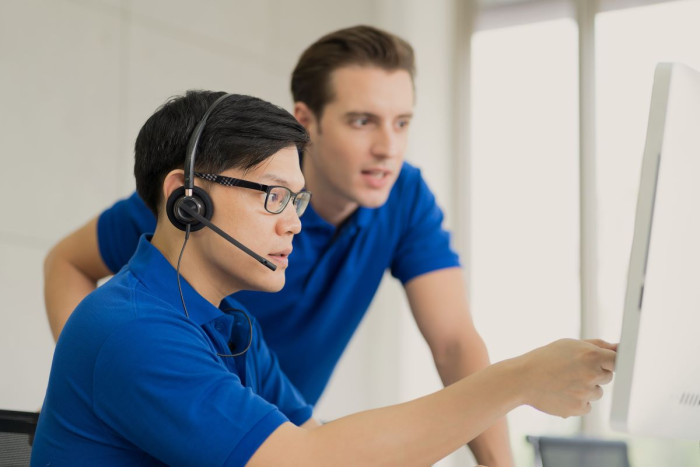 Two young men in blue shirts are in an office. One is wearing a headset and seated at a computer and pointing at it. The other is looking over his shoulder at the computer 