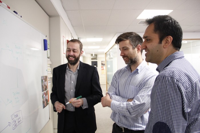 Three men stand at a whiteboard, reading a diagram and smiling