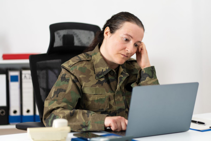 A woman in a military uniform looks at a laptop while resting her head on one hand