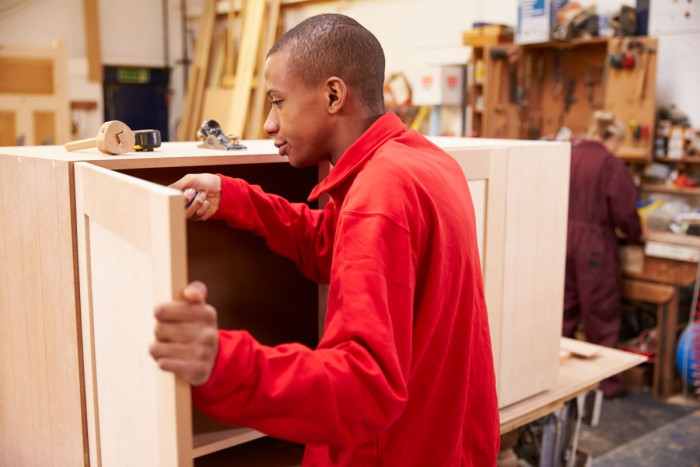 A young man using a screwdriver on a cabinet he's building in a carpentry workshop 