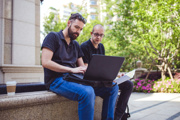 Two men in jeans and black shirts sit on a bench near a garden, looking at a laptop
