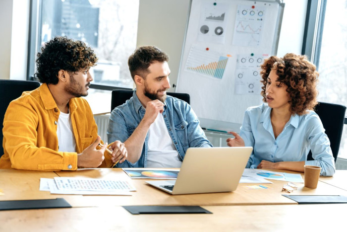 Two men and a woman in casual office clothes sit at a table in an office looking at a laptop