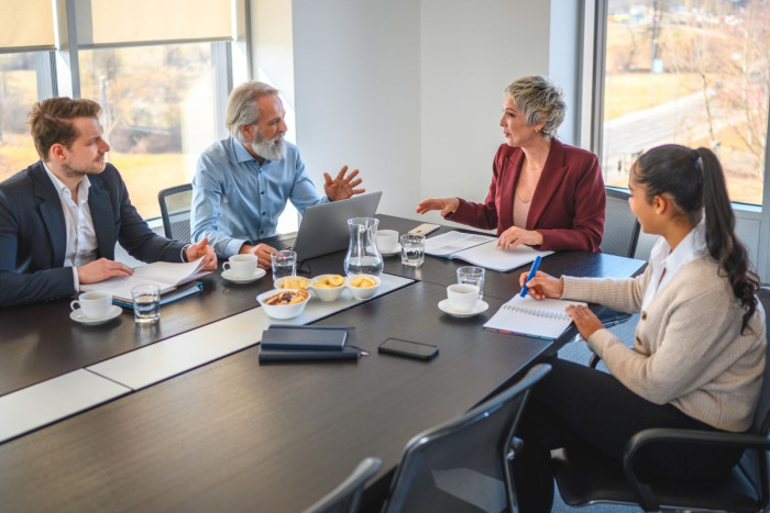 A mixed group of people in business clothes sit around a table in an office. 