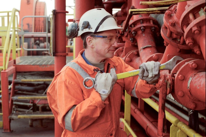 A man wearing overalls, helmet and protective gloves uses a large spanner to adjust a pipe on a ship