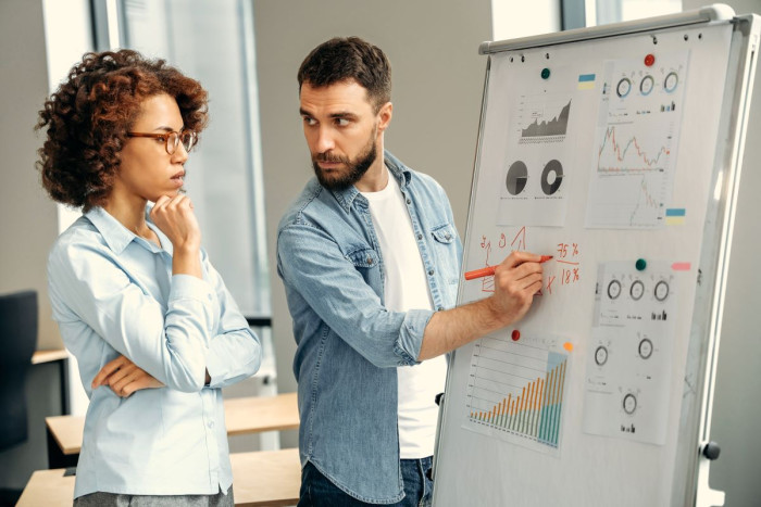 A man writes on a flip chart in an office and a woman stands looking at it with her chin in her hand