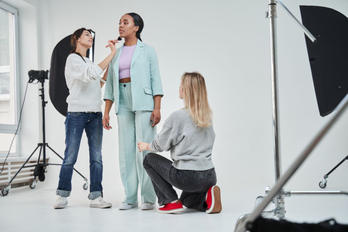 A young woman wearing a teal pants suit stands while two other people adjust her hair and clothing, with cameras and light boxes surrounding her