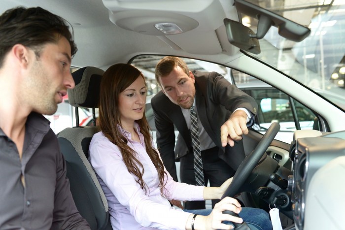 A woman sits in the driving seat of a car while a motor vehicle salesperson shows them the features of the car 