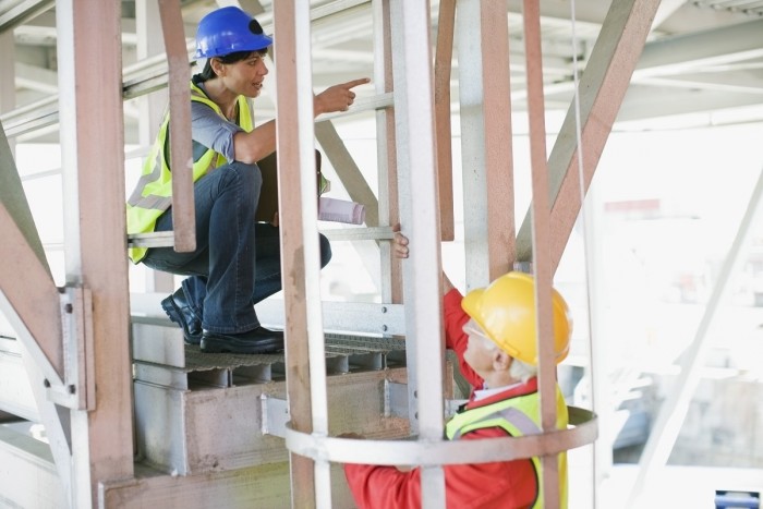 A female boat designer stands on a gangway giving directions to a male boat builder