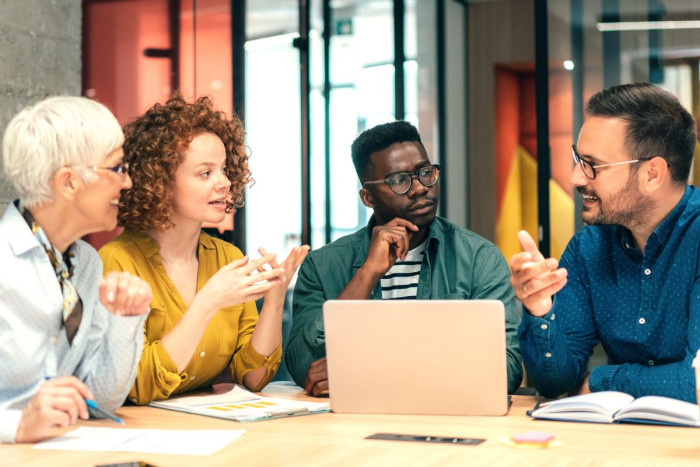 A group of people in business clothes sit a table, one man is making notes on a laptop 