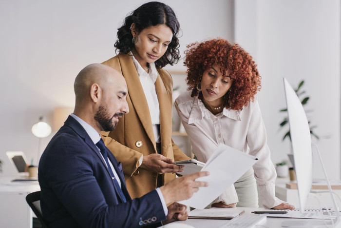 A man sits at an office desk. Two women stand beside him. They are all wearing business clothes and looking at a piece of paper he's holding 