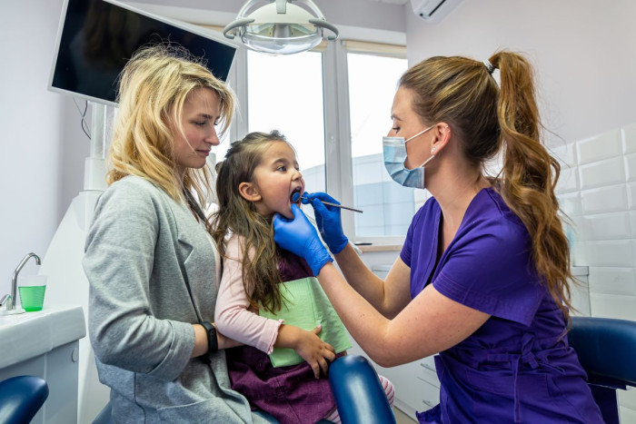 In a dental clinic, an oral health therapist wearing a mask and gloves peers into a child's mouth and inserts a specialised dental tool. The child wears protective glasses and lies  back in her mother's arms 