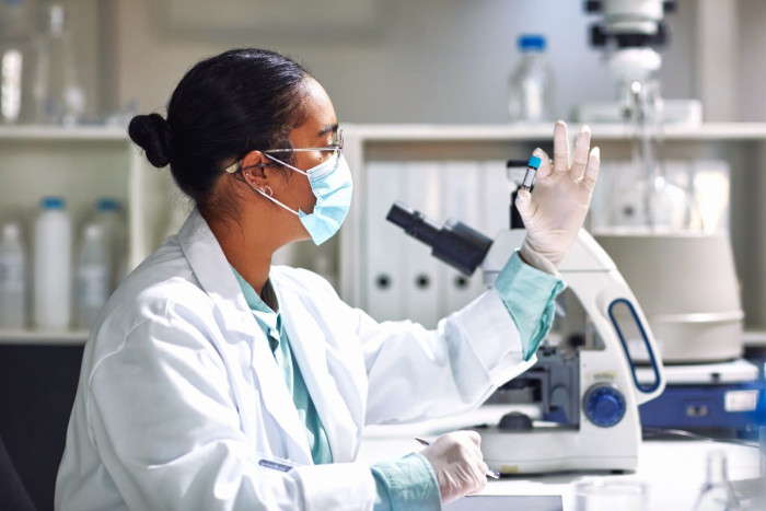 A woman wearing a lab coat and  surgical mask holds a small glass bottle with a lid up in front of a microscope