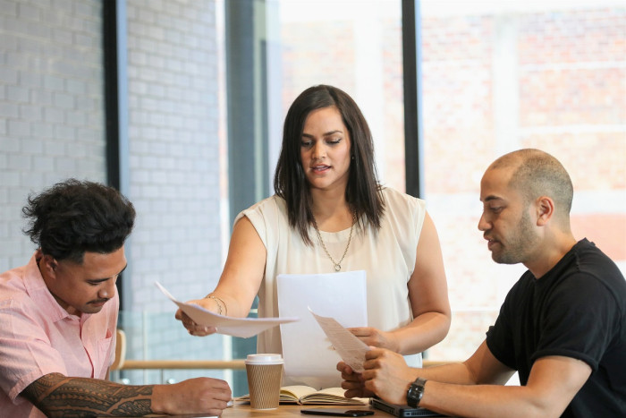 Two men sit at a table in an office. One has a tattoo on his arm. A woman stands at the head of the table handing out sheets of paper