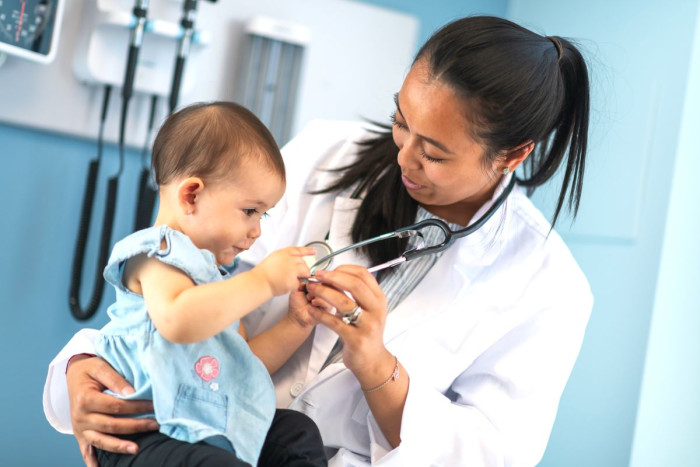 A female paediatrician wearing a white coat is holding a small child who is playing with the stethoscope