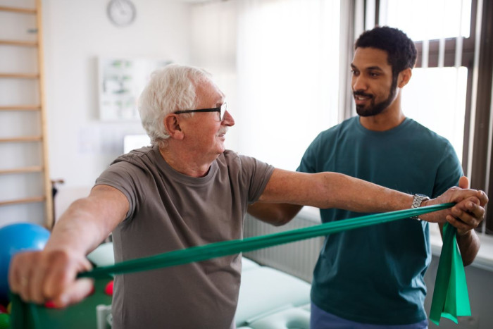 A physiotherapist stands behind an older man who is using an excerise band