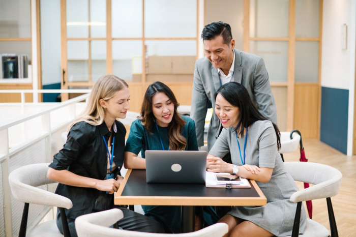 A group of women wearing office clothes and lanyards sit at a table looking at a computer. A man stands behind them looking at the computer too