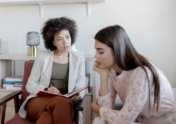 Two young women sit on chairs in an office. One is making notes in a book as she listens. The other has her chin in her hands 