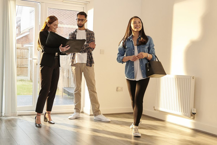 Three people stand in a sunlit empty room in a house. One holds a clipboard, one looks at it and the other looks into the room 