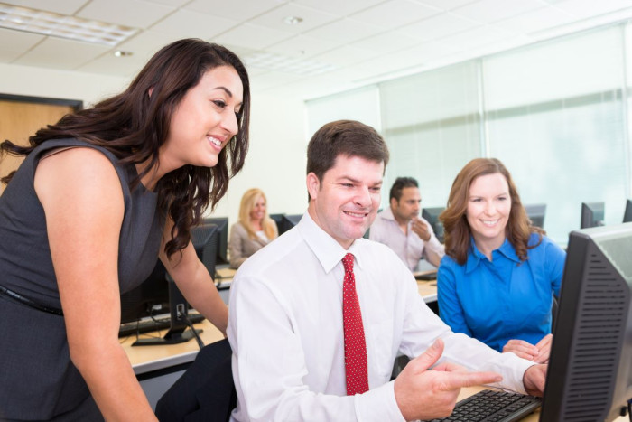 A woman leans over the shoulder of a man who is sitting at a computer in a training room. All are in business clothes 