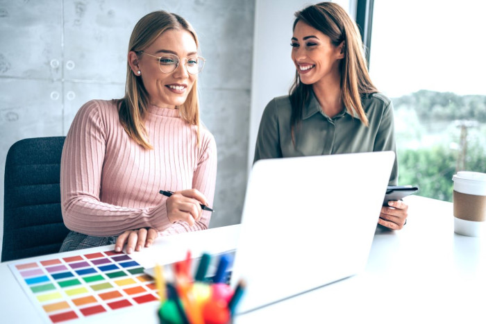 Two women sit at an office desk looking at a laptop computer with a colour chart in front of them 