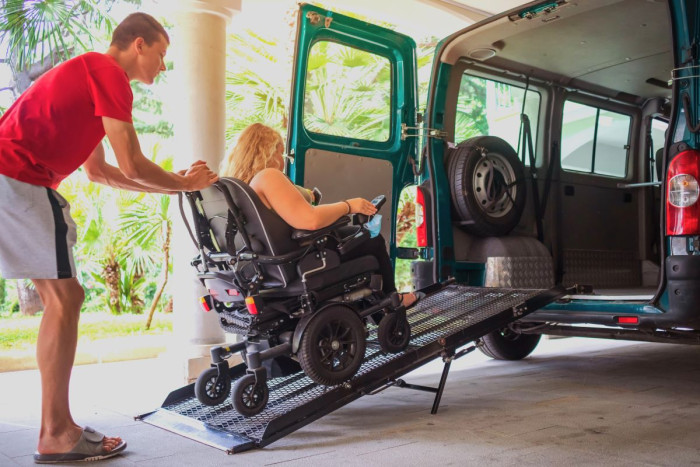 A man wearing a tee shirt and shorts steadies a woman  who is driving a motorised wheeelchair up a ramp into a van 