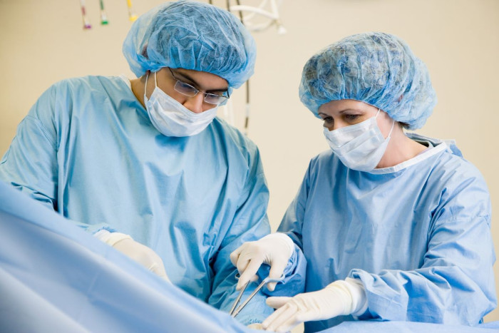 Two surgeons wearing gowns, masks, gloves and caps, stand beside a patient. One is using a surgical tool