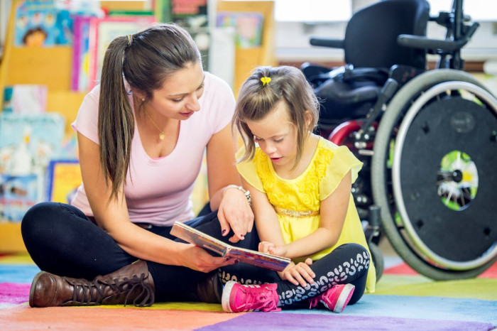 A teacher aide and a child sit on a classroom floor and look at a book, with a wheelchair in the background 
