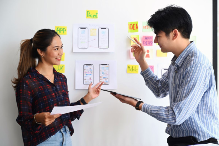 Two user experience designers stand in front of a noticeboard which is covered in drawings of app and mobile phone designs 