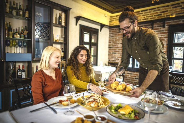 A male waiter brings another plate of food to a table in a restaurant where two women sit eating 