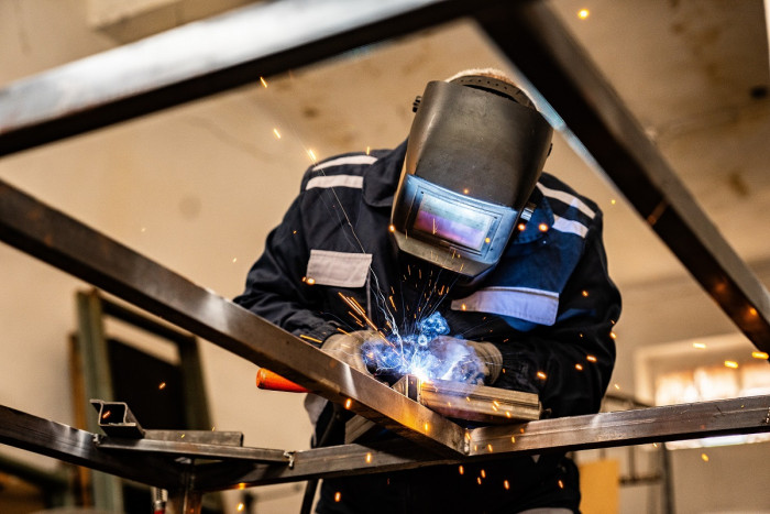 A person wears protective clothing, uses welding equipment producing bright lights and sparks