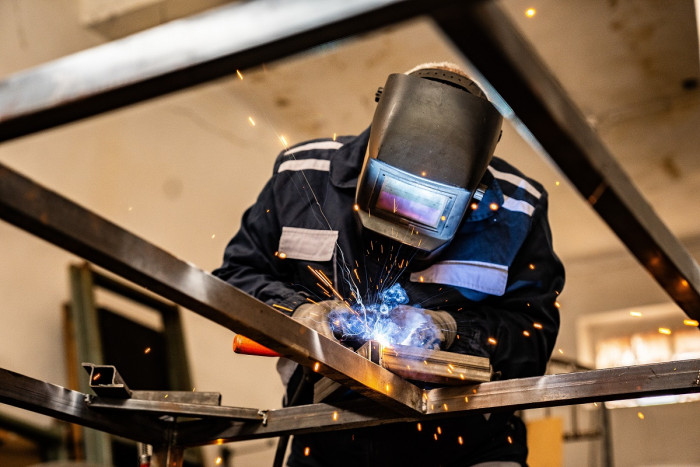 A person wears protective clothing including a thick full face mask, and uses welding equipment producing bright lights and sparks