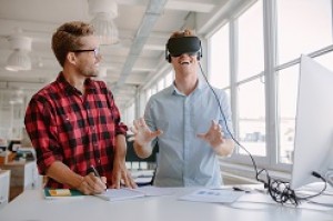 One man wears virtual reality goggles while another man stands beside him and takes notes